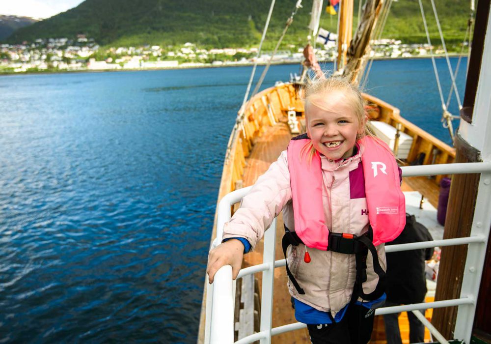 Jente i redningsvest står ved rekka om bord på Hermes II med Tromsø i bakgrunnen
